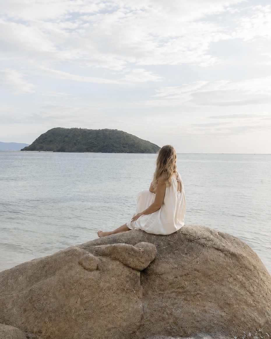 A woman in soft linen sitting on a coastal rock, gazing toward a quiet island across calm water